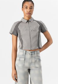 Maglietta grigia corta con zip frontale, maniche corte e piping bianco. Presenta un ricamo "Guess Originals" sul petto. I jeans in denim sono di un blu chiaro.
