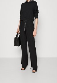 Persona con camiseta de manga larga negra y pantalones de pierna ancha con aberturas delanteras, sosteniendo un bolso negro, y llevando tacones negros abiertos.