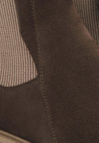 Brązowy zamszowy botek za kostkę z elastycznymi panelami bocznymi i teksturowanym materiałem, z przeszyciami podkreślającymi jego kształt i design.