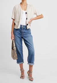 Krémová zkrácená bunda s krátkými rukávy a třemi černými knoflíky, v kombinaci s modrými zahnutejmi džínami, vzorovanou kabelkou a sandály na vysokém podpatku.