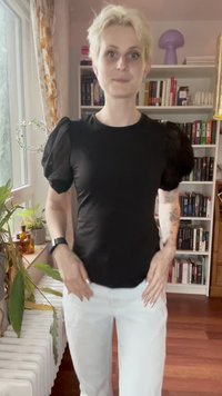 Cilvēks ar īsiem blondiem matiem, tērpies melnā puff-sleeve topā un baltās biksēs, stāv iekštelpās pie loga un grāmatu plaukta.