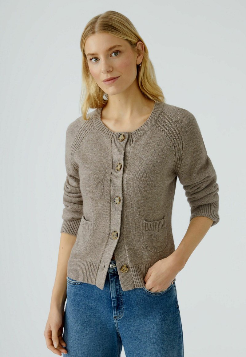 Beiger Cardigan mit rundem Ausschnitt, gerippten Bündchen, Knopfverschluss und zwei vorderen Taschen, aus weichem Strickmaterial.