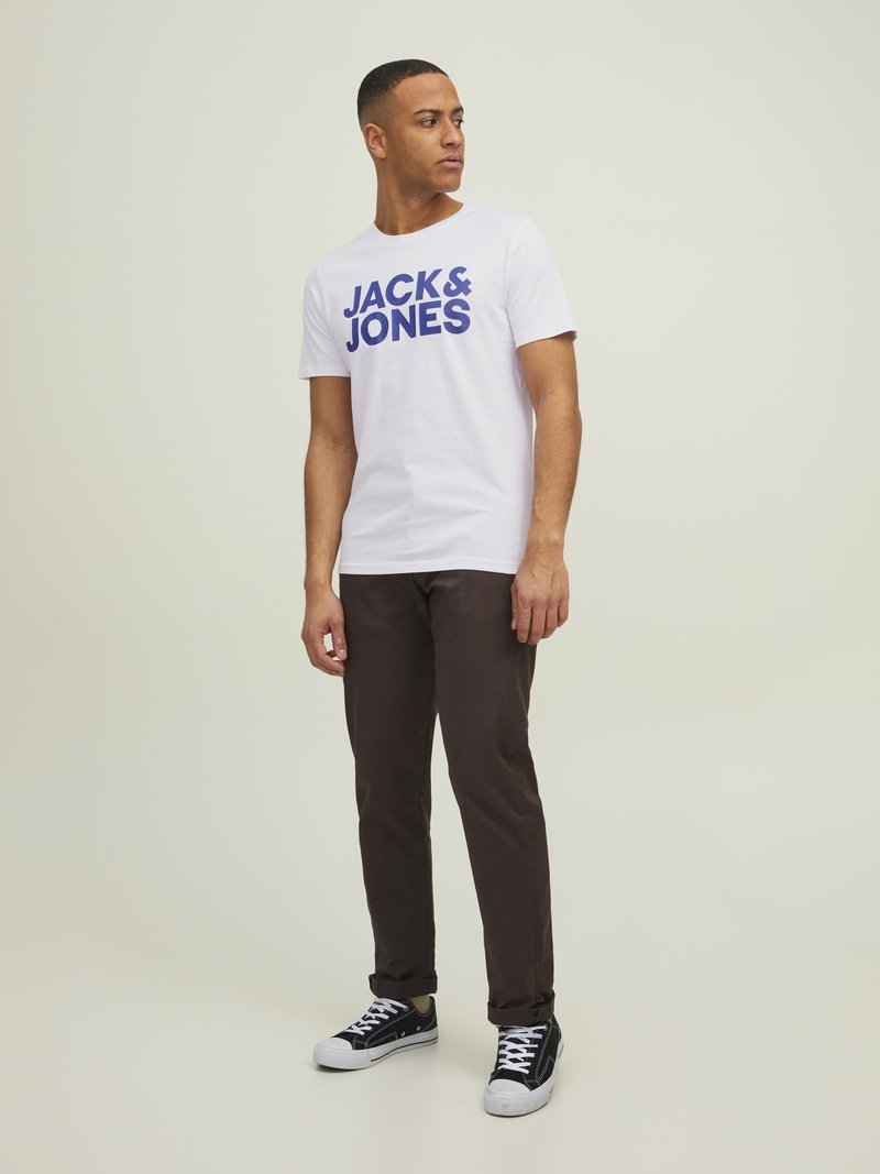 Bijela majica s velikim plavim natpisom "JACK & JONES", kontrastne smeđe hlače i crne tenisice s bijelim potplatima, stojeći uz svijetlu pozadinu.
