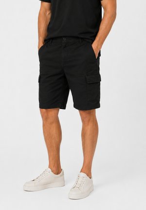 Mężczyzna w czarnych szortach cargo, czarnym t-shircie i białych sneakersach stoi z rękami w kieszeniach na tle jasnego, gładkiego tła.
