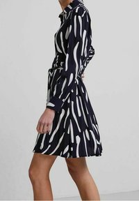 Donna vestita con un abito a maniche lunghe nero e bianco a fantasia, con gonna a pieghe, mentre cammina su uno sfondo grigio chiaro uniforme.