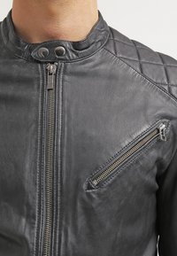 Nahaufnahme einer Person, die eine schwarze Lederjacke mit einem Frontreißverschluss, gesteppten Schulterdetails und einer diagonalen, mit Reißverschluss versehenen Brusttasche trägt.