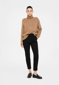 Donna in piedi che indossa un maglione a collo alto color cammello, pantaloni neri a sigaretta, mocassini neri e orecchini a cerchio dorati, su uno sfondo bianco semplice.