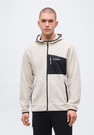 Giovane uomo che indossa una giacca in pile beige Columbia con tasche nere con cerniera sul petto e sui lati, in piedi davanti a uno sfondo chiaro e uniforme.