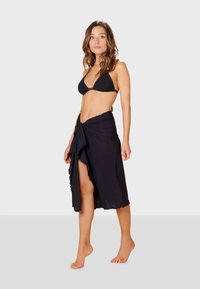 Musta sarong, jossa on sulava kuvio, rypytetty helma ja sidottu vyötärö. Pukeutuneena mustan bikinin päälle, esittelee kevyttä kangasta ja tekstuuria.