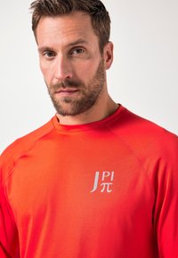 Punainen urheilullinen pitkähihainen paita, jossa pyöreä pääntie ja hopeinen logo vasemmalla puolella, jossa lukee "J Pi π". Sileä tekstuuri, istuva malli.