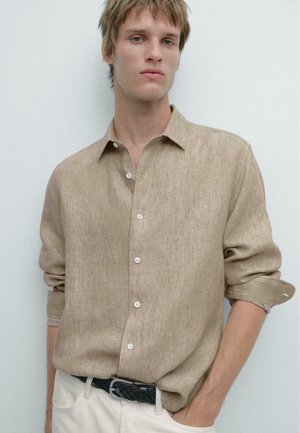 Giovane uomo con le maniche arrotolate che indossa una camicia beige di lino abbottonata e infilata in pantaloni chiari con una cintura intrecciata nera, in piedi contro un muro bianco.