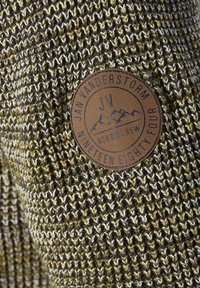 Strickstoff mit schwarzen, weißen und olivfarbenen Fäden, ausgestattet mit einem runden Lederpatch mit der Aufschrift "Jan Vanderstorm, JV, Nor Crew, Nineteen Eighty Four."