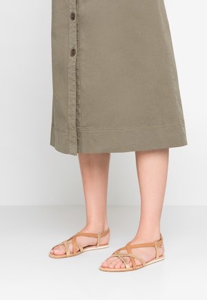 Jambes inférieures portant des sandales plates couleur tan avec des brides croisées et une jupe kaki boutonnée tombant sous les genoux sur fond blanc.