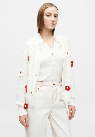 Hvit strikket cardigan med røde og kremfargede blomsterapplikasjoner, med kabelstrikkdesign, knappelukking og ribbestrikkede mansjetter. Brukt over en lys topp.