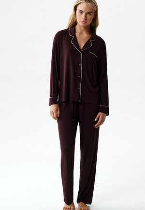 Kobieta ubrana w ciemnofioletową, zapinaną na guziki, długorękawową piżamę z dopasowanymi spodniami z białym lamówką, stojąca boso na białym tle.