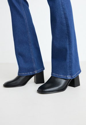 Jambes portant un jean bleu et des bottines en cuir noir à talons blocs, debout sur un sol clair.