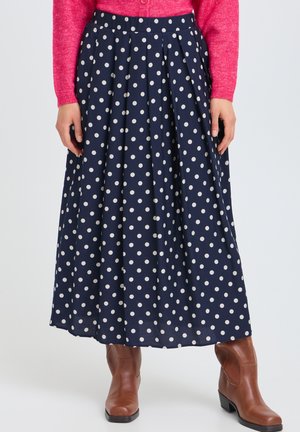 Personne portant une jupe longue bleu marine à pois blancs, un pull rose et des bottines marron, debout droit.