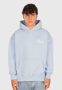 Junger Mann mit kurzem, lockigem Haar, der einen hellblauen Hoodie mit Fronttasche und dem Text "Dropsee Street Game" trägt, kombiniert mit hellen Jeans.