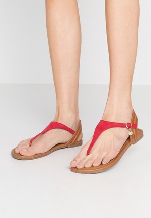 Pies con sandalias planas rojas y marrones con diseño de tira en T y un pequeño charm metálico redondo en la correa del tobillo.