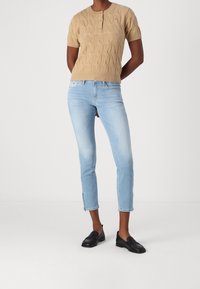 Pull court en tricot beige à col Henley; jeans ajustés bleu clair avec des zips aux chevilles; mocassins noirs.