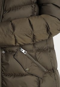 Giacca piumino verde oliva con texture trapuntata, una tasca con zip sul davanti e hardware della cerniera argento a contrasto.