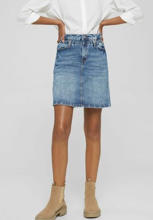 Kvinde iført en blå denimnederdel, hvid skjorte med opkørede ærmer og beige ankelstøvler, stående med hænderne på hofterne mod en grå baggrund.