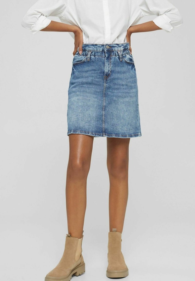 Femme portant une jupe en jean bleu, une chemise blanche aux manches retroussées et des bottines beiges, debout les mains sur les hanches devant un fond gris.