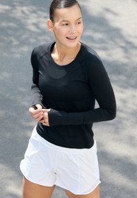Crna dugačka majica s dugim rukavima od sportske tkanine s rebrastom teksturom, uparena s bijelim šortsem. Majica ima rupice za palčeve i okrugli izrez.