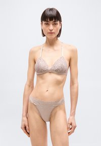 Γυναίκα που ποζάρει με μπεζ δαντελένιο σουτιέν με λεπτές τιράντες και ταίριαστα εσώρουχα με δαντέλα στο περίγραμμα, σε απλό φωτεινό φόντο.