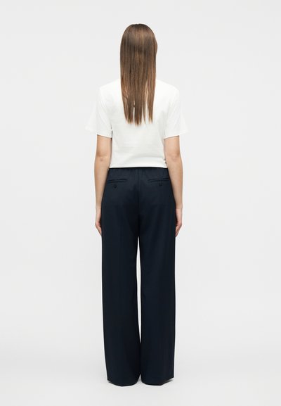 Γυναίκα με μακριά καστανά μαλλιά που φοράει λευκό κοντομάνικο τοπ και άνετο μαύρο παντελόνι, στεκόμενη με την πλάτη προς τα εμάς μπροστά σε απλό φόντο.