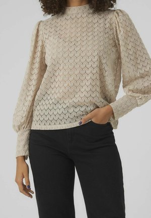 Beżowa koronkowa bluzka z teksturowanym wzorem fal, z wysokim kołnierzem i bufkami na długich rękawach, zestawiona z czarnymi dopasowanymi spodniami.