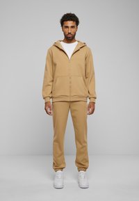 Beige Zip-Hoodie mit einer Vordertasche, kombiniert mit passenden Jogginghosen. Das Modell trägt ein weißes T-Shirt und weiße Sneaker. Glatte Textur.