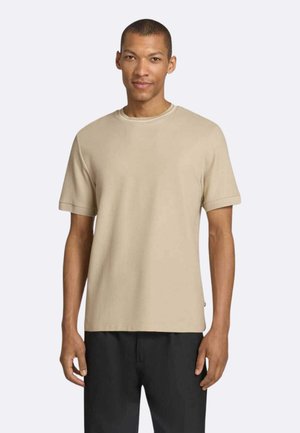Uomo che indossa una maglietta beige a maniche corte e pantaloni neri, in piedi davanti a uno sfondo chiaro e uniforme.