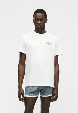 Άνδρας που φοράει άσπρο t-shirt Moschino και μπλε τζιν σορτς, στέκεται μπροστά σε ένα απλό ανοιχτό γκρι φόντο.