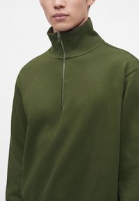 Homme portant un pull à demi-fermeture éclair de couleur vert olive avec le zip partiellement fermé au niveau du col montant.