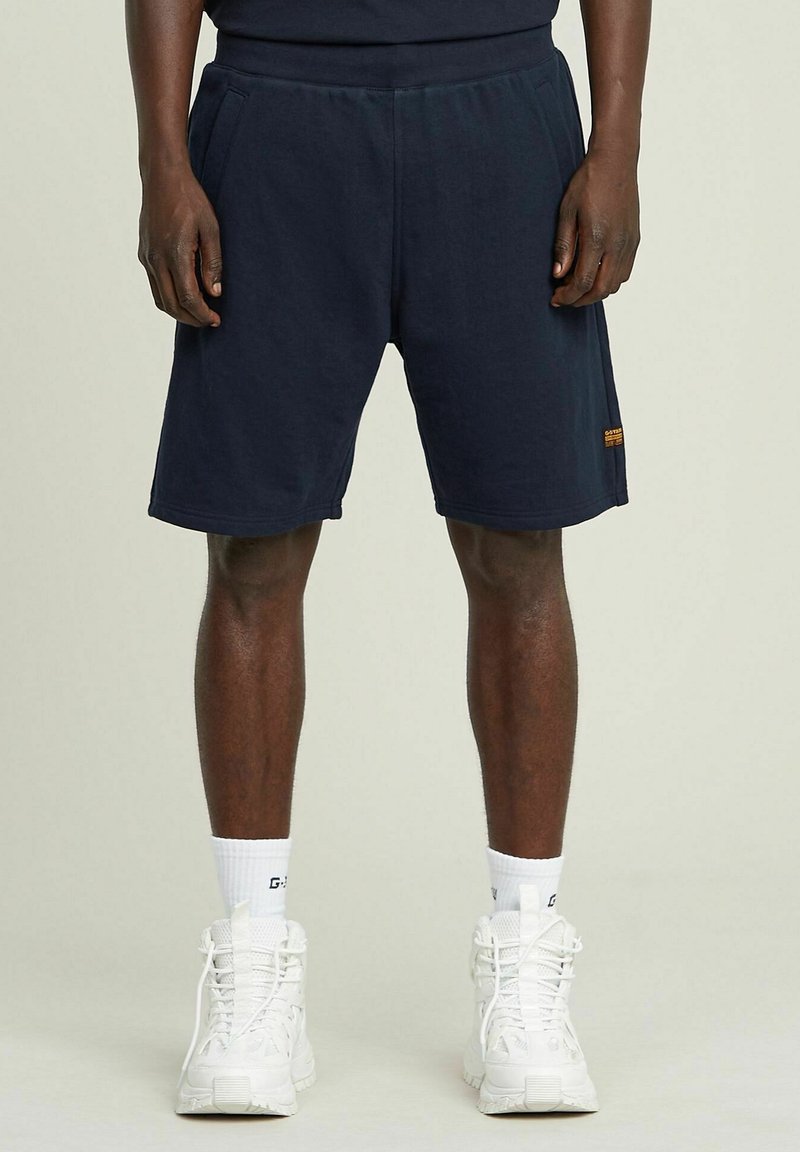 Shorts in cotone blu navy con una vita elastica, due tasche laterali e una piccola etichetta arancione vicino all'orlo.