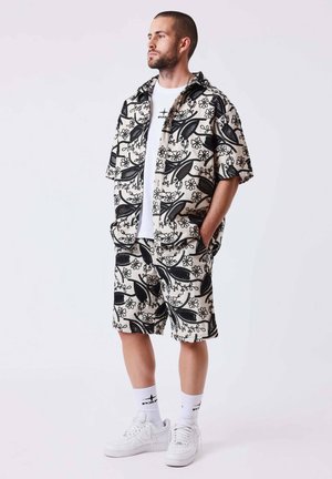 Mann steht mit den Händen in den Taschen und trägt ein passendes schwarz-weißes Blumenmuster-Hemd und Shorts, ein weißes T-Shirt mit Grafik, Socken und Turnschuhe.