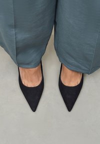 Crne antilop cipele sa špicastim vrhom na bosim nogama, uparene s laganim, lepršavim tirkiznim hlačama koje sežu do pola lista, na neutralnoj pozadini.