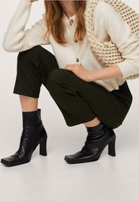 Zwarte leren enkellaarzen met een vierkante neus en een stevige hak. Gecombineerd met olijfgroene broek en een crèmekleurige cardigan, aangevuld met een getextureerde tas.