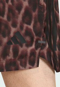 Ruskeat shortsit, joissa on leopardikuvio ja musta Adidas-logo. Valmistettu teksturoidusta kankaasta, jossa sivuhalkiot liikkuvuutta varten.