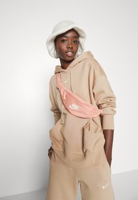 Riñonera rosa de Nike con cierre con cremallera, llevada sobre un sudadera beige y pantalones deportivos a juego. El modelo lleva un sombrero de cubo blanco de felpa.