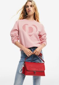 Felpa rosa con design a forma di cuore e lettera 'D', abbinata a jeans in denim. Una borsa a tracolla in pelle rossa presenta testo in rilievo e hardware in argento.