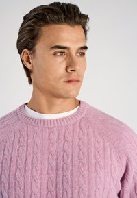 Ljusrosa kabelstickad tröja med rund halsringning, med ribbad krage och axelsömmar. Bärs över en vit t-shirt.