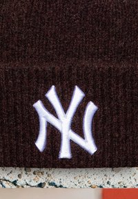Tamsiai rudos spalvos mezginys su baltu išsiuvinėtu tarpusavyje susijungiančiu "NY" logotipu, esančiu centre, arti apatinio audinio krašto.
