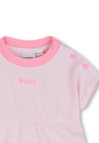 Robe côtelée rose clair avec un col rond, ornée d'un texte de logo rose et de boutons-pression sur une épaule. Tissu léger avec une texture douce.