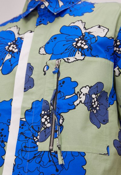 Camicia floreale con grandi fiori blu su uno sfondo verde, dotata di una tasca sul petto con zip, colletto a punte e una striscia bianca.
