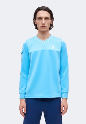 Homme aux cheveux foncés mi-longs portant un haut de sport turquoise à manches longues col en V et un pantalon bleu marine, debout devant un fond clair.