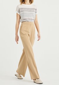 Donna che cammina indossando pantaloni a vita alta beige a gamba larga, mocassini crema e un top in maglina a maniche corte con motivi in bianco e nero.