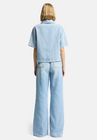 Lys blå denim cropped jakke med krave og korte ærmer, parret med brede lys blå denimbukser. Baghånden viser lomme detaljer.