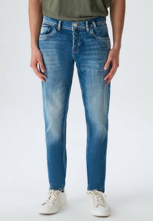 Homme portant un jean slim bleu délavé et des baskets blanches, debout les mains détendues le long du corps sur un fond uni.